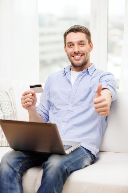 concepto de tecnología, hogar y estilo de vida - hombre sonriente trabajando con laptop y tarjeta de crédito en casa y mostrando los pulgares hacia arriba