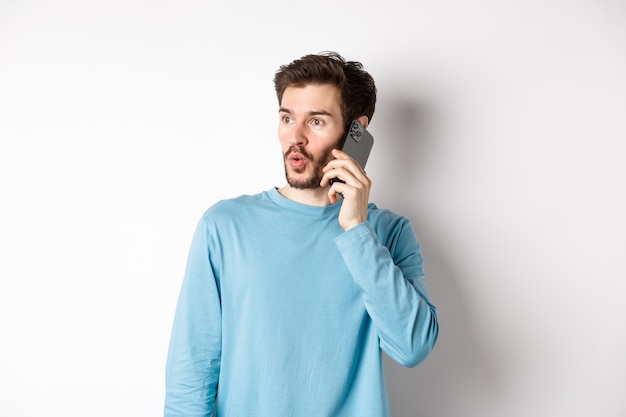 Concepto de tecnología. Joven intrigado mirando a la izquierda impresionado, hablando por teléfono móvil, llamando a alguien, de pie sobre fondo blanco.