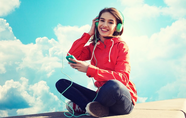 concepto de tecnología, música y personas: mujer joven sonriente o adolescente con smartphone y auriculares escuchando música al aire libre sobre el cielo azul y el fondo de las nubes