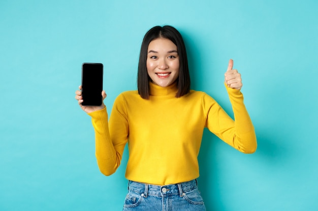 Concepto de tecnología y personas. Chica asiática alegre en suéter amarillo que muestra la pantalla del teléfono inteligente en blanco y los pulgares hacia arriba, demuestra la oferta en línea, de pie sobre fondo azul.