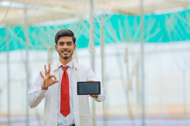 Concepto de tecnología y personas, joven agrónomo indio mostrando tableta o teléfono inteligente en invernadero