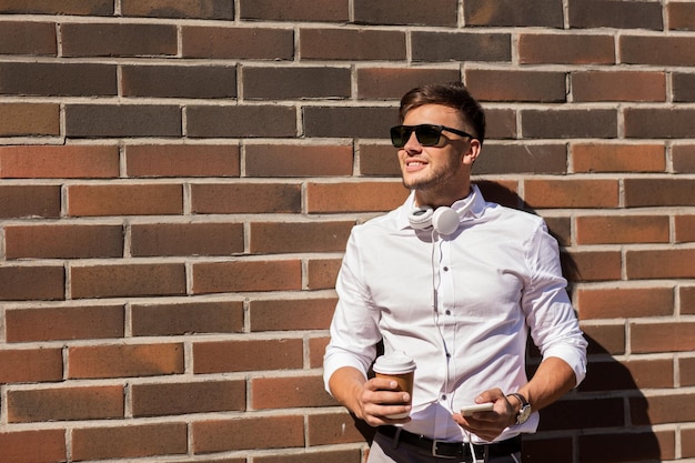 concepto de tecnología y personas - joven sonriente feliz con gafas de sol con teléfono inteligente y taza de café en la calle de la ciudad