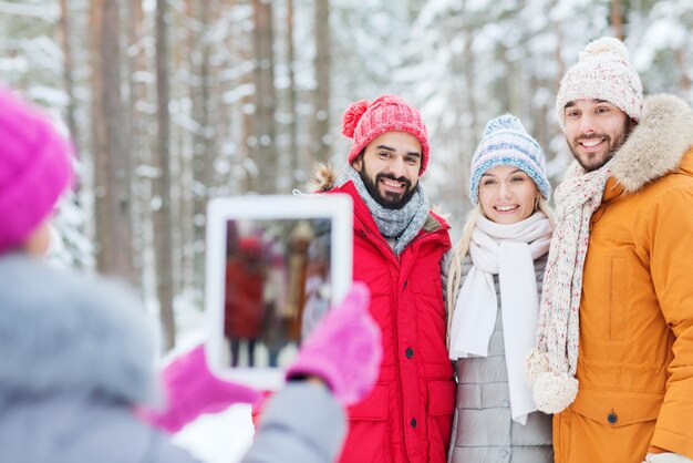 concepto de tecnología, temporada, amistad y personas - grupo de hombres y mujeres sonrientes tomando fotos con una computadora de tablet pc en el bosque de invierno