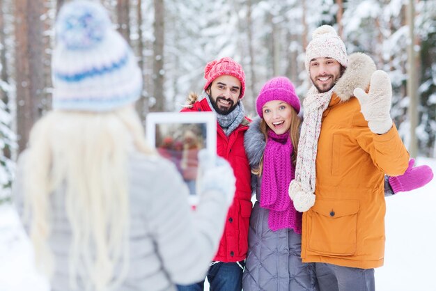 concepto de tecnología, temporada, amistad y personas - grupo de hombres y mujeres sonrientes tomando fotos con una computadora de tablet pc en el bosque de invierno