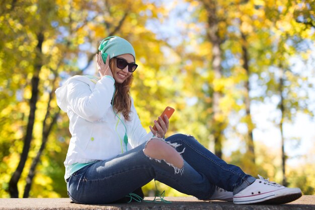 Foto concepto de tecnología, temporada y personas - mujer joven sonriente o adolescente con smartphone y auriculares escuchando música sobre el fondo del parque de otoño