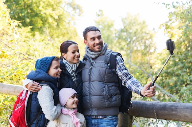 concepto de tecnología, viajes, turismo, caminatas y personas - familia feliz con mochilas tomando fotos con un smartphone en selfie stick y caminatas