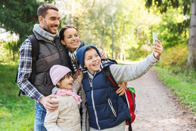 Foto concepto de tecnología, viajes, turismo, caminatas y personas - familia feliz con mochilas tomando selfie por teléfono inteligente y caminatas