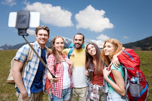 Foto concepto de tecnología, viajes, turismo, caminatas y personas - grupo de amigos sonrientes caminando con mochilas tomando fotos por teléfono inteligente en selfie stick sobre fondo natural