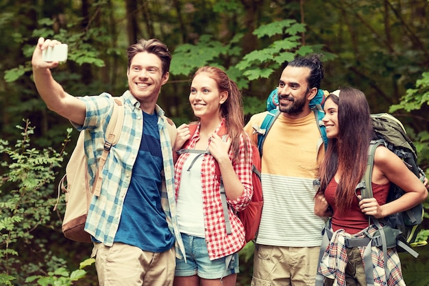 concepto de tecnología, viajes, turismo, caminatas y personas - grupo de amigos sonrientes caminando con mochilas tomando selfie por teléfono inteligente en el bosque