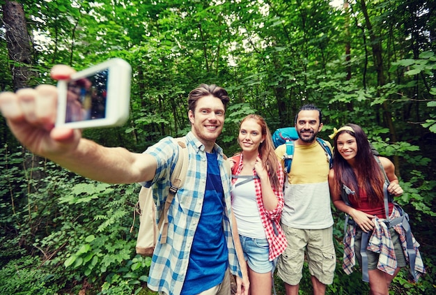 Foto concepto de tecnología, viajes, turismo, caminatas y personas - grupo de amigos sonrientes caminando con mochilas tomando selfie por teléfono inteligente en el bosque