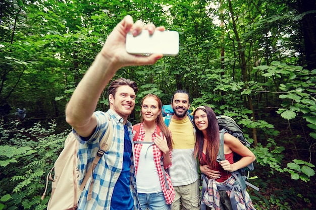 concepto de tecnología, viajes, turismo, caminatas y personas - grupo de amigos sonrientes caminando con mochilas tomando selfie por teléfono inteligente en el bosque