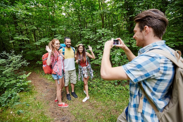 concepto de tecnología, viajes, turismo, caminatas y personas - grupo de amigos sonrientes con mochilas tomando fotos por teléfono inteligente y agitando las manos en el bosque
