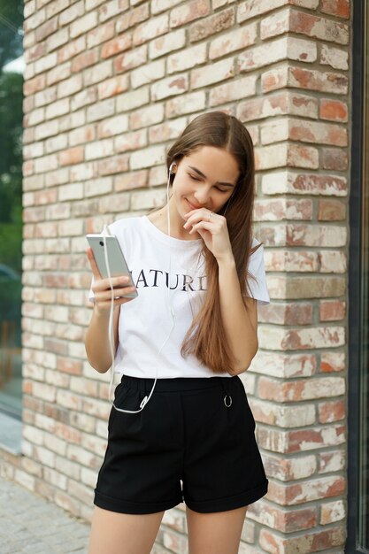 Concepto de tecnologías, emociones, personas, música, belleza, moda y estilo de vida: mujer joven con auriculares colgando de su teléfono móvil mientras camina en una calle urbana, vista desde una altura