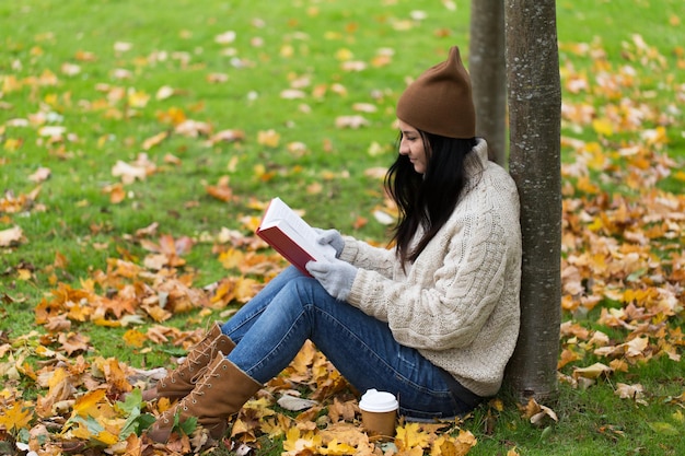 concepto de temporada, literatura, educación y personas - mujer joven leyendo un libro y bebiendo café de una taza de papel en el parque de otoño