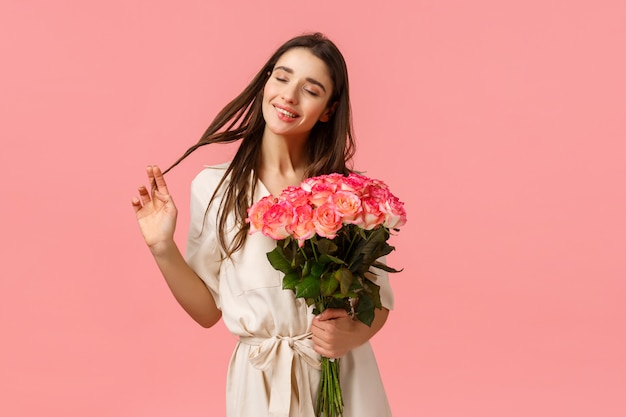 Concepto de ternura, deleite y día de San Valentín. Encantadora, encantadora y sensual mujer morena vestida, enrollando el cabello sobre los ojos felizmente soñando, recibiendo flores, sosteniendo rosas