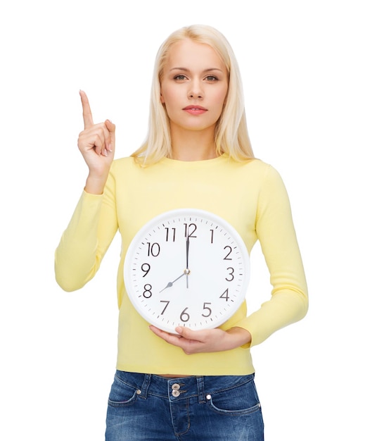 concepto de tiempo, educación y personas - mujer joven seria con reloj de pared que muestra 8 y dedo hacia arriba