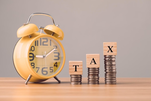 Foto concepto de tiempo de pago de impuestos tax escribir en bloques de madera dispuestos encima de la pila de monedas y reloj amarillo en el escritorio de la oficina con pared gris