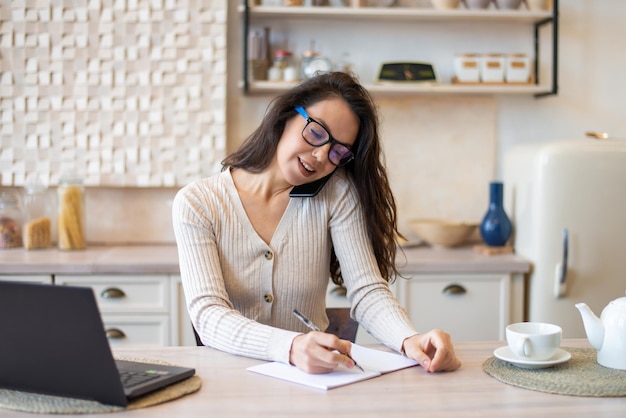 Concepto de trabajo independiente señora confiada hablando por teléfono inteligente y tomando notas sentado en la cocina