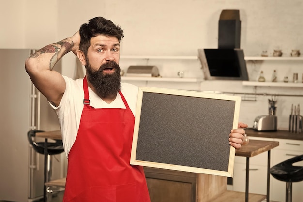 Concepto de trucos de cocina Formas inteligentes de organizar la cocina Cocinar con espacio de copia de pizarra en blanco Consejos secretos Información útil Hombre barbudo hipster delantal rojo de pie en la cocina Tienda de muebles de cocina