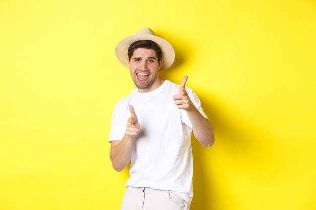 Concepto de turismo y vacaciones. Feliz chico cool apuntando con el dedo hacia ti, de pie sobre fondo amarillo