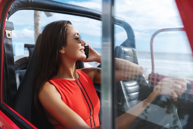 Concepto de turismo y vacaciones tropicales Hermosa mujer en traje de baño conduciendo un coche todoterreno en viajes de verano a la playa hablando por teléfono y sonriendo