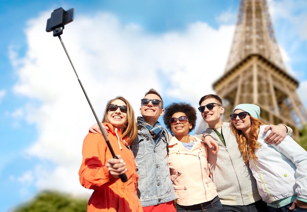 concepto de turismo, viajes, personas, ocio y tecnología - grupo de amigos adolescentes sonrientes tomando selfie con smartphone y monopod sobre el fondo de la torre eiffel de parís