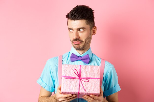 Concepto de vacaciones y celebración. Chico sorprendido mirando a un lado con la cara encogida, sosteniendo una pequeña caja de regalo, de pie sobre un fondo rosa.