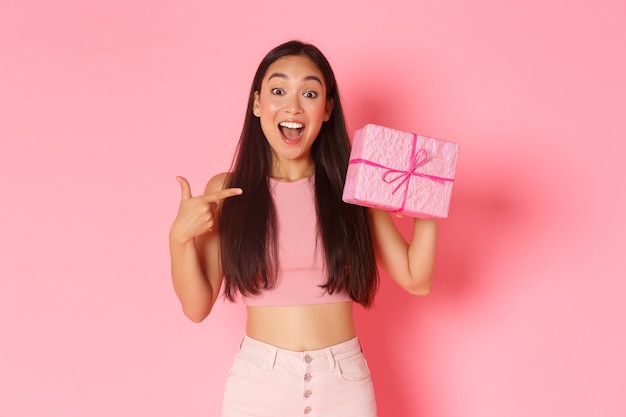 Concepto de vacaciones, celebración y estilo de vida. Chica asiática sorprendida y emocionada, feliz adivinando qué hay dentro de la caja de regalo, apuntando al presente y sonriendo optimista, de pie sobre una pared rosa.