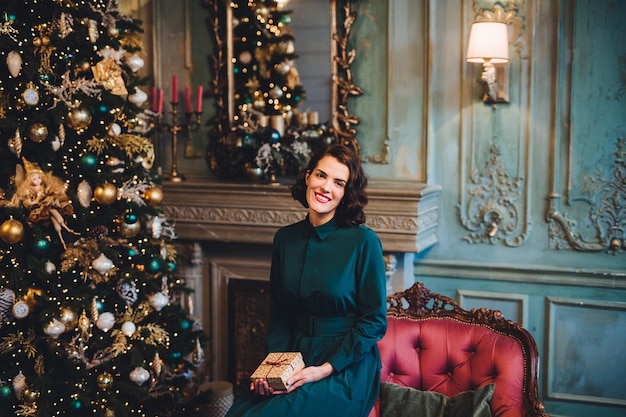 Concepto de vacaciones de celebración de personas Una mujer hermosa y sonriente usa un vestido agradable se sienta cerca del árbol de Navidad en la habitación y tiene un presente feliz de celebrar su fiesta favorita Feliz Navidad y Año Nuevo