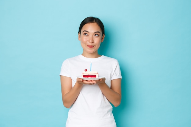 Concepto de vacaciones, estilo de vida y celebración. Retrato de niña asiática sonriente soñadora en camiseta blanca mirando pensativo mientras sostiene un trozo de pastel de cumpleaños, pidiendo deseos en la vela encendida.