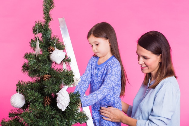 Concepto de vacaciones, familia y navidad - madre e hija decorando el árbol de navidad en rosa