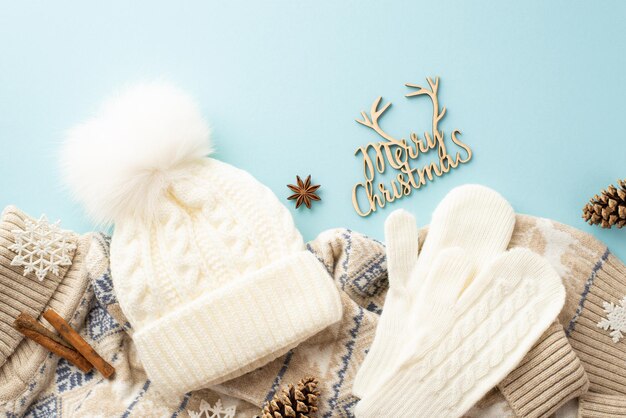 Concepto de vacaciones de invierno Foto de vista superior de feliz navidad texto de madera guantes blancos bobble sombrero tejido suéter copos de nieve piñas anís y canela sobre fondo azul pastel aislado