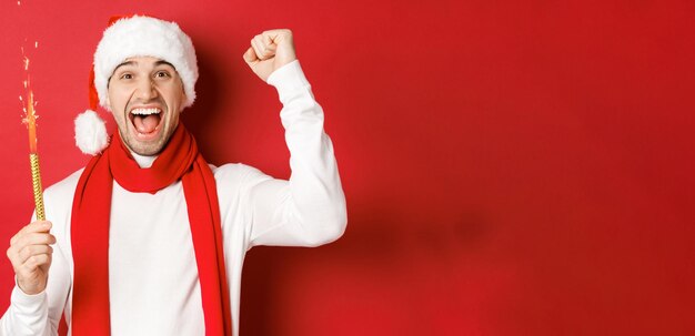 Concepto de vacaciones de invierno de navidad y celebración Hombre guapo celebrando el año nuevo y divirtiéndose sosteniendo bengala y sonriendo con sombrero de santa de pie sobre fondo rojo