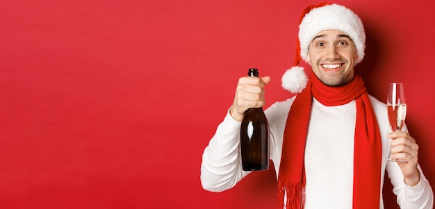 Concepto de vacaciones de invierno Navidad y estilo de vida Primer plano de un hombre guapo alegre sosteniendo una botella de champán y un vaso haciendo brindis por el año nuevo y celebrando el fondo rojo