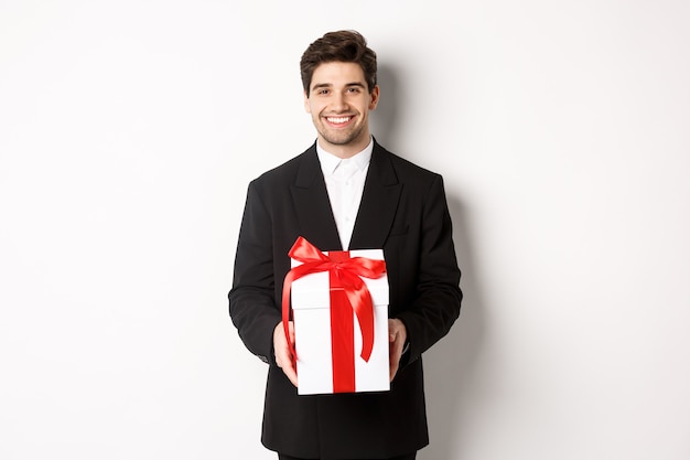 Concepto de vacaciones de Navidad, celebración y estilo de vida. Hombre guapo en traje negro, tiene presente romántico, sosteniendo un regalo en una caja y sonriendo, de pie contra el fondo blanco.