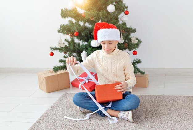 Foto concepto de vacaciones, navidad, infancia y personas - sonriente feliz muchacho adolescente con sombrero de santa abre caja de regalo sobre fondo de árbol de navidad.