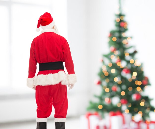 concepto de vacaciones y personas - hombre disfrazado de santa claus desde atrás sobre la sala de estar y el fondo del árbol de navidad