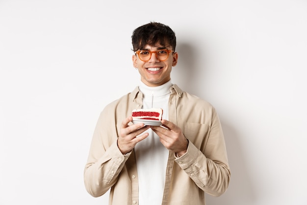 Concepto de vacaciones y personas reales. Chico hipster feliz en vasos celebrando un cumpleaños, pidiendo un deseo en el b-day cake con vela y sonriendo, de pie en la pared blanca.