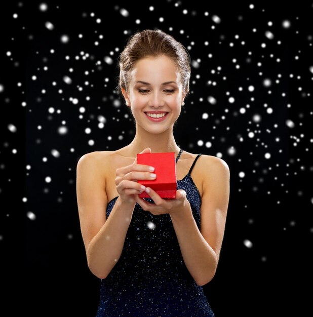 concepto de vacaciones, regalos, lujo y felicidad - mujer sonriente vestida sosteniendo una caja de regalo roja sobre fondo negro nevado