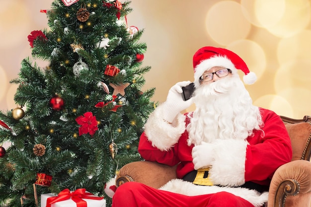 concepto de vacaciones, tecnología y personas - hombre disfrazado de santa claus con smartphone, regalos y árbol de navidad sobre fondo de luces beige