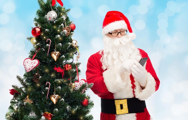 concepto de vacaciones, tecnología y personas - hombre disfrazado de santa claus con teléfono inteligente y árbol de navidad sobre fondo de luces azules