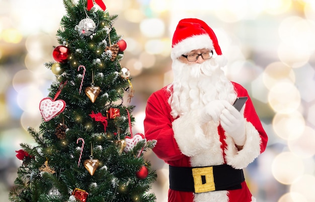 concepto de vacaciones, tecnología y personas - hombre disfrazado de santa claus con teléfono inteligente y árbol de navidad sobre fondo de luces azules