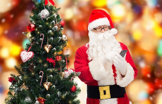 concepto de vacaciones, tecnología y personas - hombre disfrazado de santa claus con teléfono inteligente y árbol de navidad sobre fondo de luces rojas