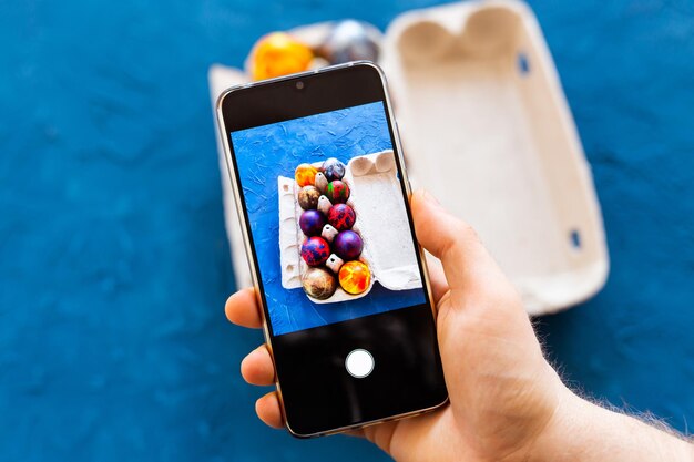 Concepto de vacaciones, tradición, tecnología y personas - cerca de las manos del hombre con el teléfono inteligente tomando fotografías de huevos de pascua de colores.