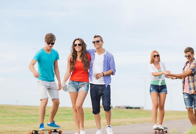 concepto de vacaciones, vacaciones, amor y amistad - grupo de adolescentes sonrientes caminando y montando en patinetas al aire libre