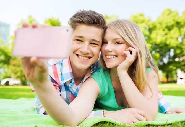 concepto de vacaciones, vacaciones, tecnología y amistad - pareja sonriente tirada en una manta y haciendo selfie con smartphone en el parque