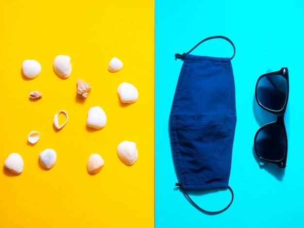 Concepto de vacaciones de verano y viajes en las condiciones de la pandemia covid-19. Gafas, conchas y máscara sobre una mesa amarilla y azul. Lay Flat, copia espacio.