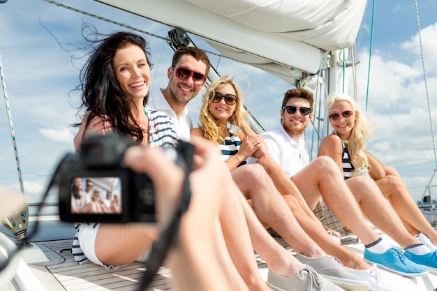 concepto de vacaciones, viajes, mar, amistad y personas - amigos sonrientes sentados en la cubierta del yate y fotografiando