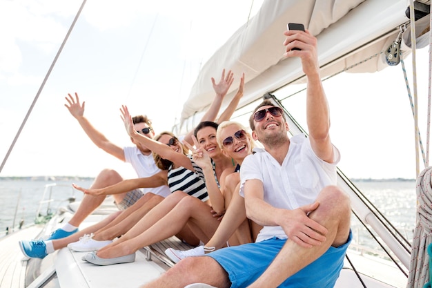 concepto de vacaciones, viajes, mar, amistad y personas - amigos sonrientes sentados en la cubierta del yate y haciendo selfie