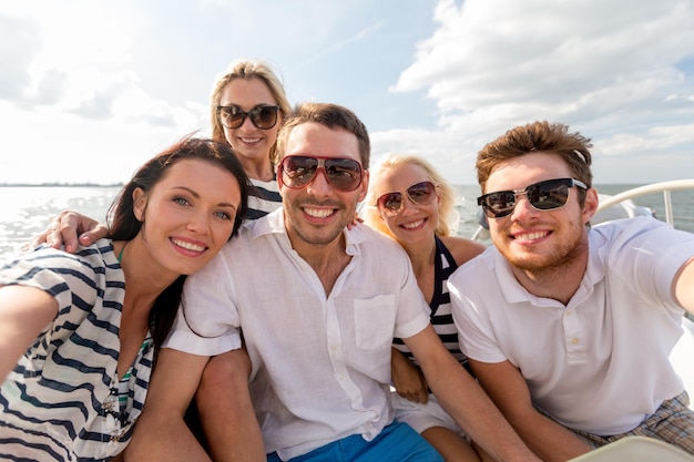 concepto de vacaciones, viajes, mar, amistad y personas - amigos sonrientes sentados en la cubierta del yate y haciendo selfie